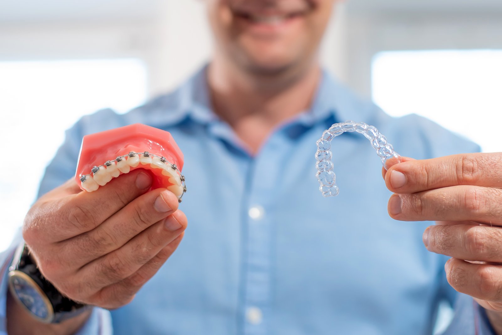 Adult holding braces and a clear aligner to compare Invisalign vs. braces